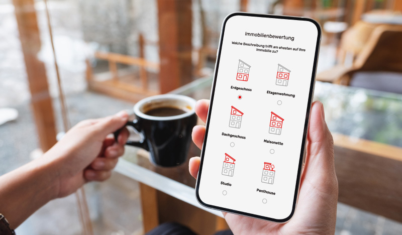 Eine Person hält ein Smartphone in der Hand, auf dem die Online-Maske von Leben am Bodensee zur Immobilienbewertung geöffnet ist. Auf dem Bildschirm sind verschiedene Auswahlmöglichkeiten wie Erdgeschoss, Etagenwohnung, Dachgeschoss, Maisonette, Studio und Penthouse zu sehen. Im Hintergrund steht eine Tasse Kaffee auf einem Tisch in einem Café.